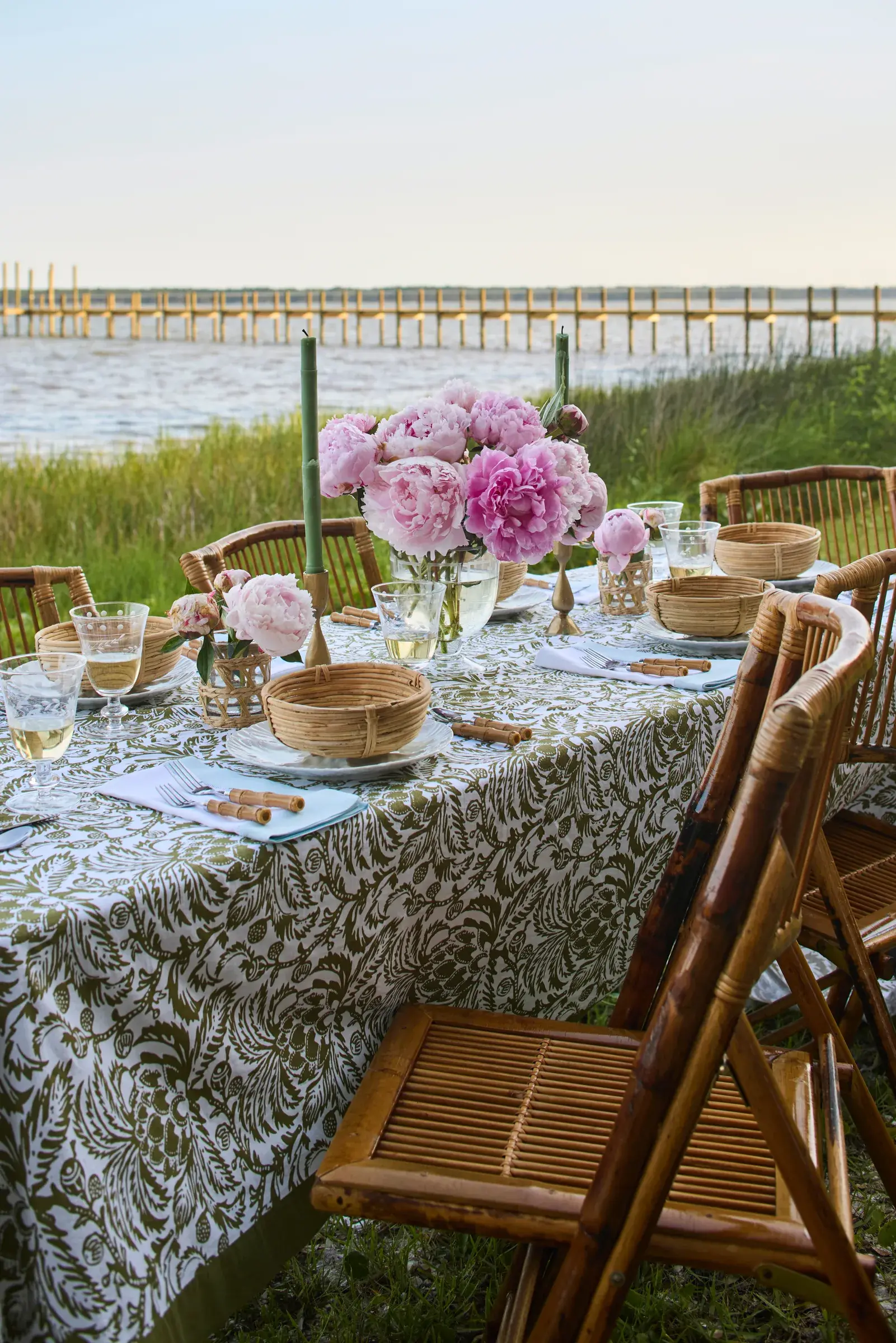 A long table at the marsh edge set with peonies and bamboo chairs.